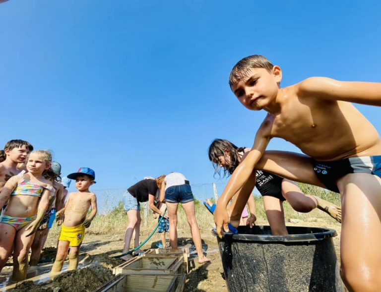 Copii în costume de baie lângă o găleată mare cu lut, la Chirpici pentru cei mici