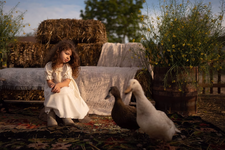 Little girl sitting on straw bales, watching two ducks in the yard of Ferma Lipovenesc, Jurilovca