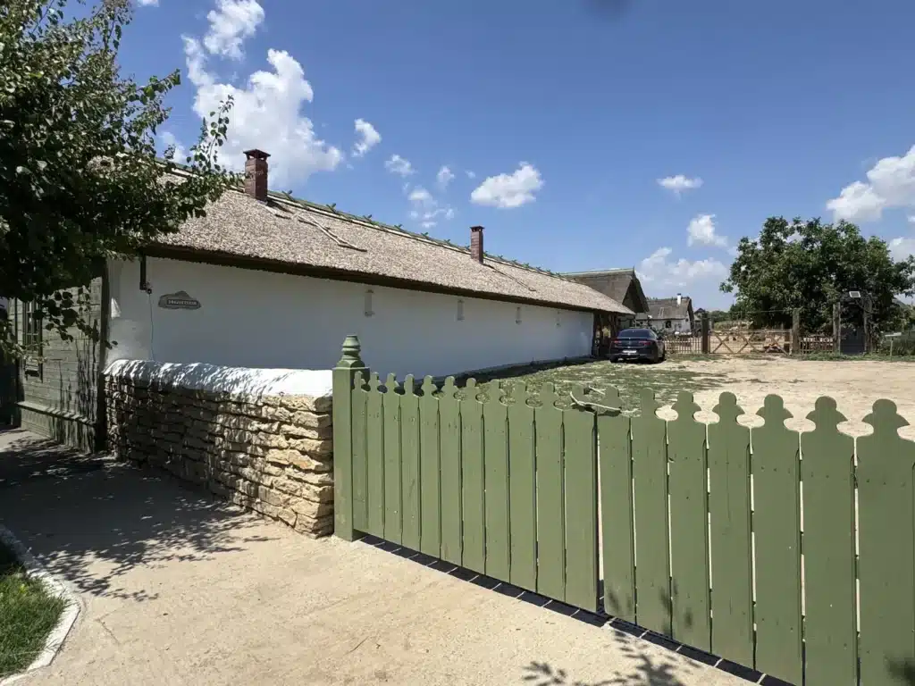 Casa veche din 1893 de la Ferma Lipovenesc după restaurare, lungă și albă, cu acoperiș de stuf, văzută de la poarta verde a curții