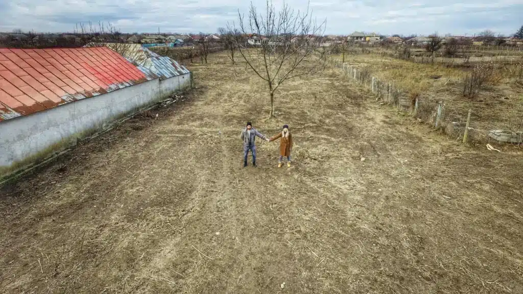 Curtea goală din Jurilovca văzută din dronă în 2020, cu casa veche acoperită cu tablă roșie și Marian și Daniela Sterea ținându-se de mână